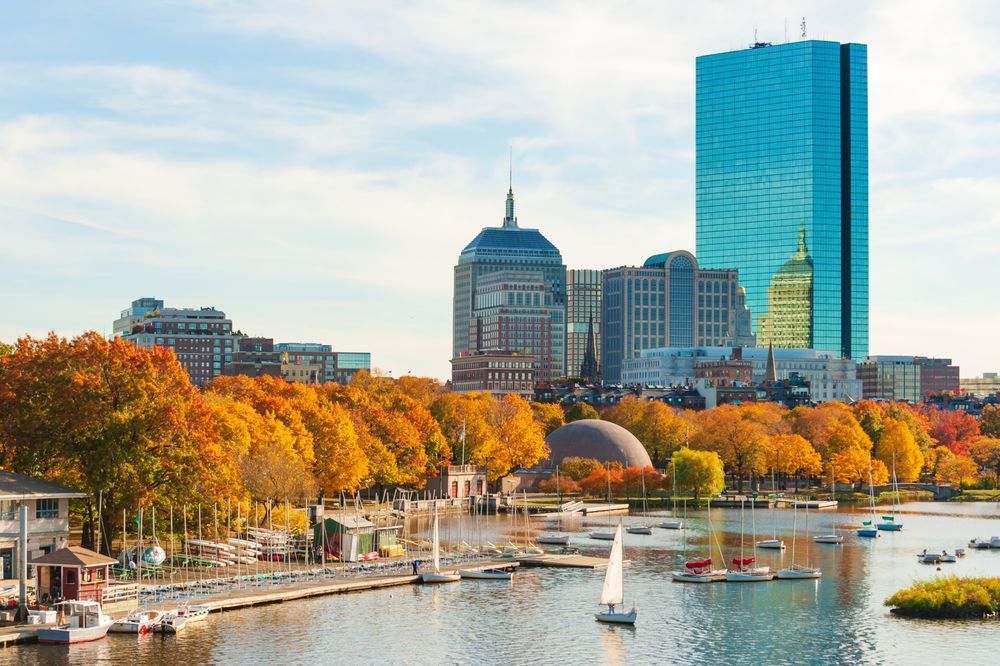 A riverside cityscape in New England, USA during autumn, with vibrant orange and yellow trees along the water, sailboats docked and sailing on a calm river, and a mix of modern and historic buildings in the background under a clear blue sky.
