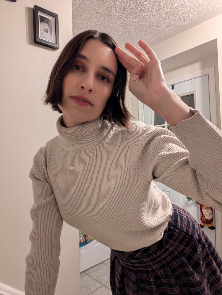 A girl poses wearing a plaid skirt and beige turtleneck 
