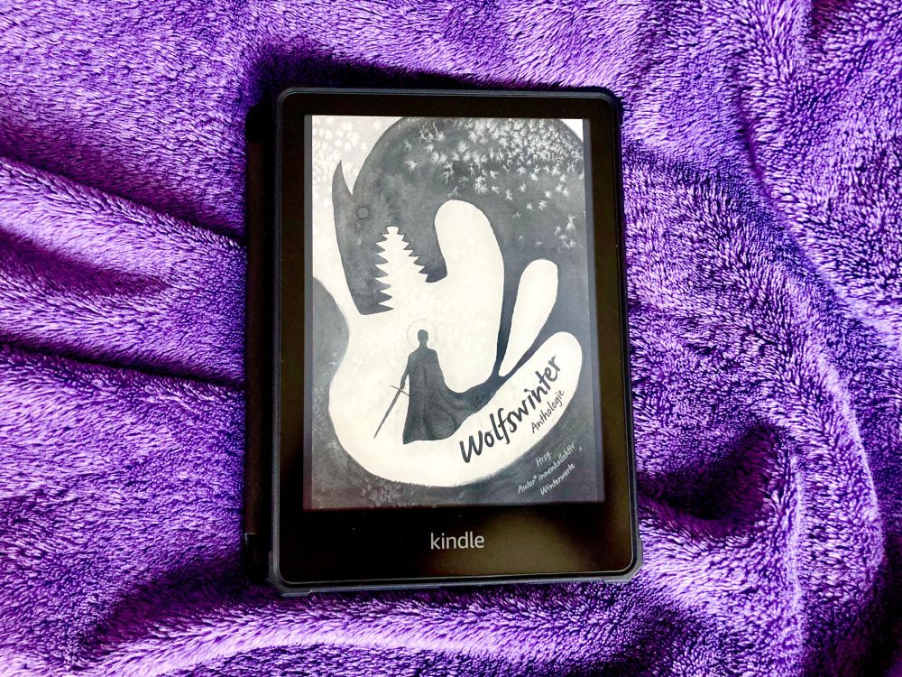 "Wolfswinter" vom Autor*innenkollektiv Winterworte auf einem Kindle E-Reader. Auf dem gezeichneten Cover ist die Silhouette einer menschlichen Gestalt mit einem Schwert in der Hand zu sehen. Sie trägt einen Umhang, der am Saum in eine Wolfsgestalt übergeht, die sich mit aufgerissenem Maul und spitzen Zähnen über den Menschen beugt. In der rechten unteren Ecke steht in Handschrift: "Wolfswinter. Anthologie. Hrsg. Autor*innenkollektiv Winterworte".