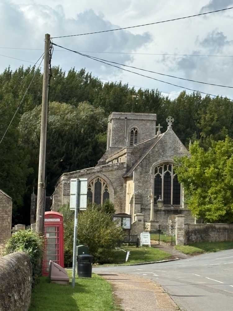 Tansor church nestled in front of a high bank of trees. View from the east of a little tower and a small stone church. Just to the left of the church are a red phone box and a telegraph pole 