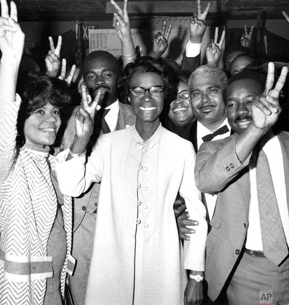 Shirley Chisholm smiling in the center of a group of joyful people, all raising their hands with peace signs at an event.
