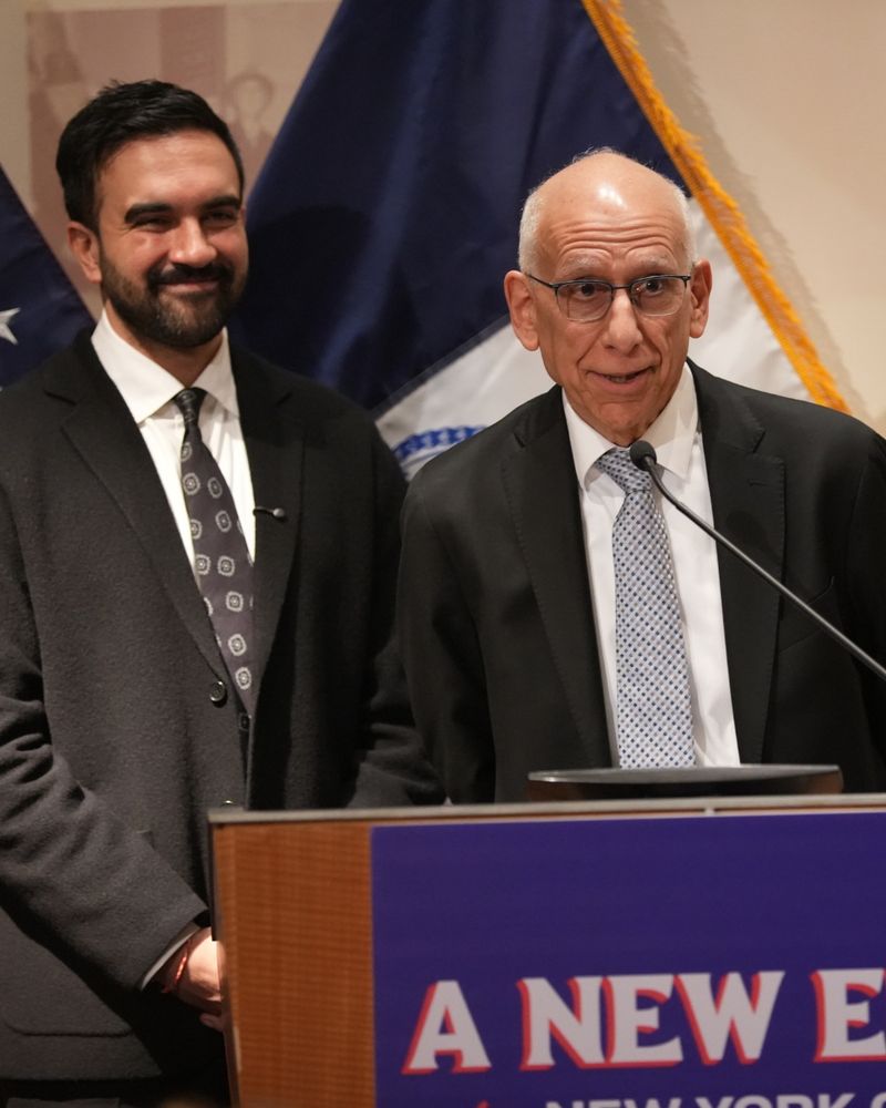 Mayor-elect Zohran Mamdani and Dean Fuleihan stand at a podium during an event. The podium has a blue sign with bold red and white text that reads: “A NEW ERA for NEW YORK CITY” 