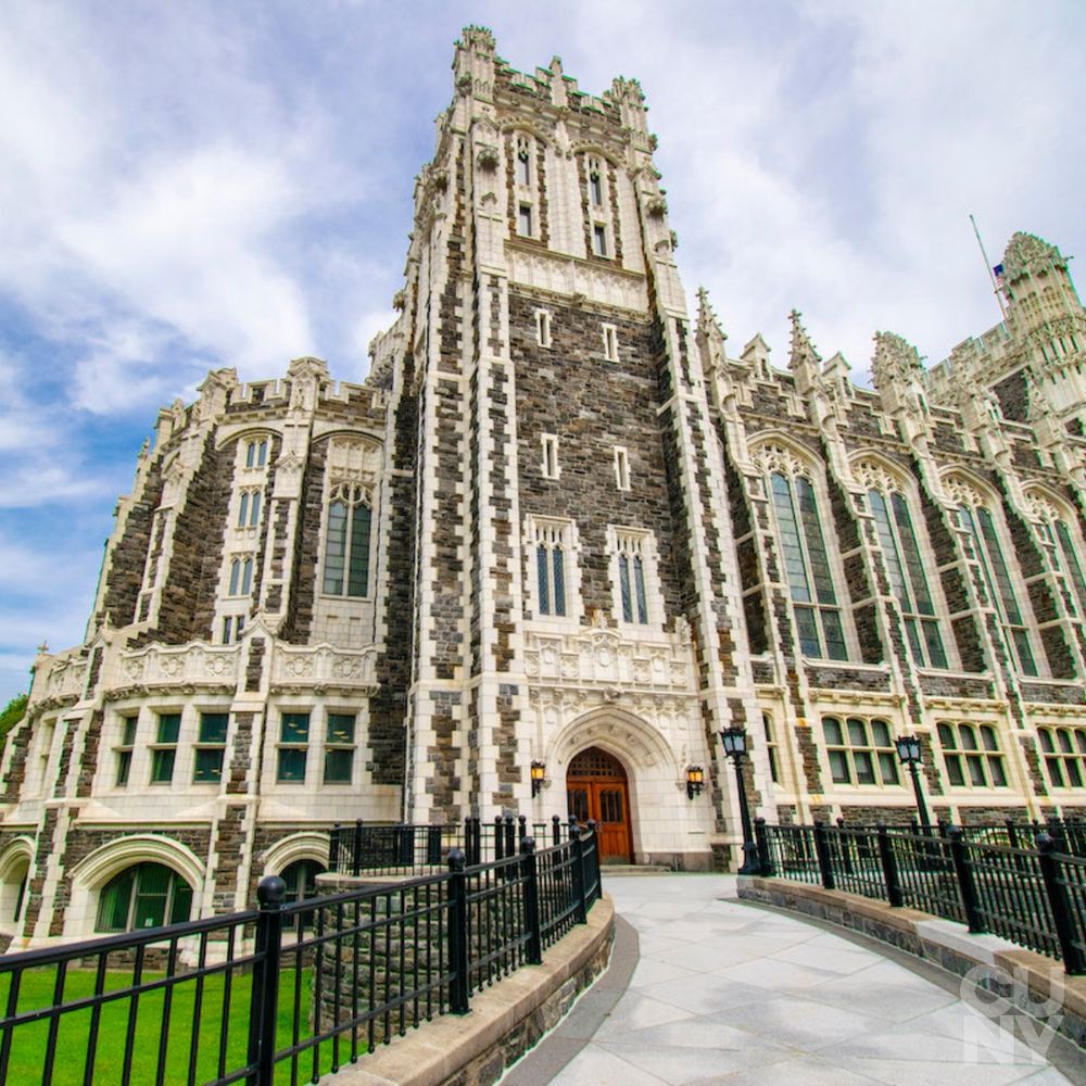 Gothic-style building with intricate stonework, tall narrow windows, and a central tower flanked by two smaller towers. The facade features contrasting dark and light stones, with a pathway leading to an ornate arched entrance. Partly cloudy sky enhances the dramatic architecture