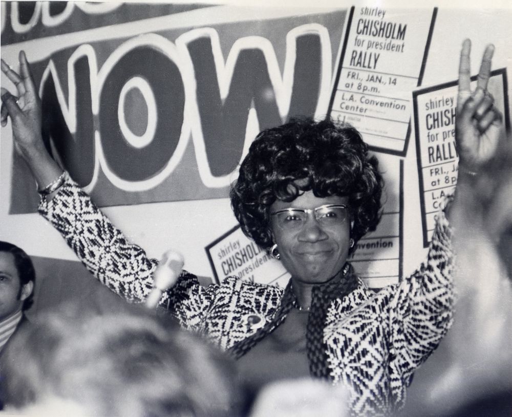 Shirley Chisholm giving a speech at a rally, with arms raised in front of a 'WOW' sign and other campaign posters.