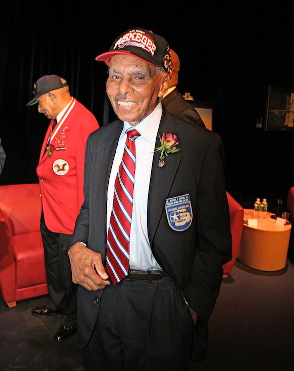 Roscoe Brown wearing a Tuskegee Airmen cap and a red blazer with medals, standing and smiling at an indoor event. 