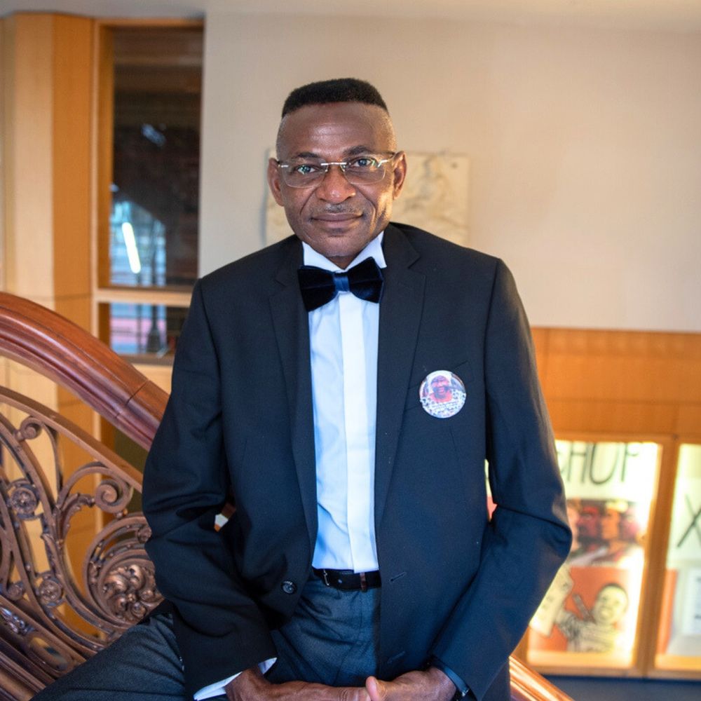 Dr. Eugene Mananga wearing a formal black suit with a bow tie and a badge, sitting on a staircase railing, smiling in a well-lit room.
