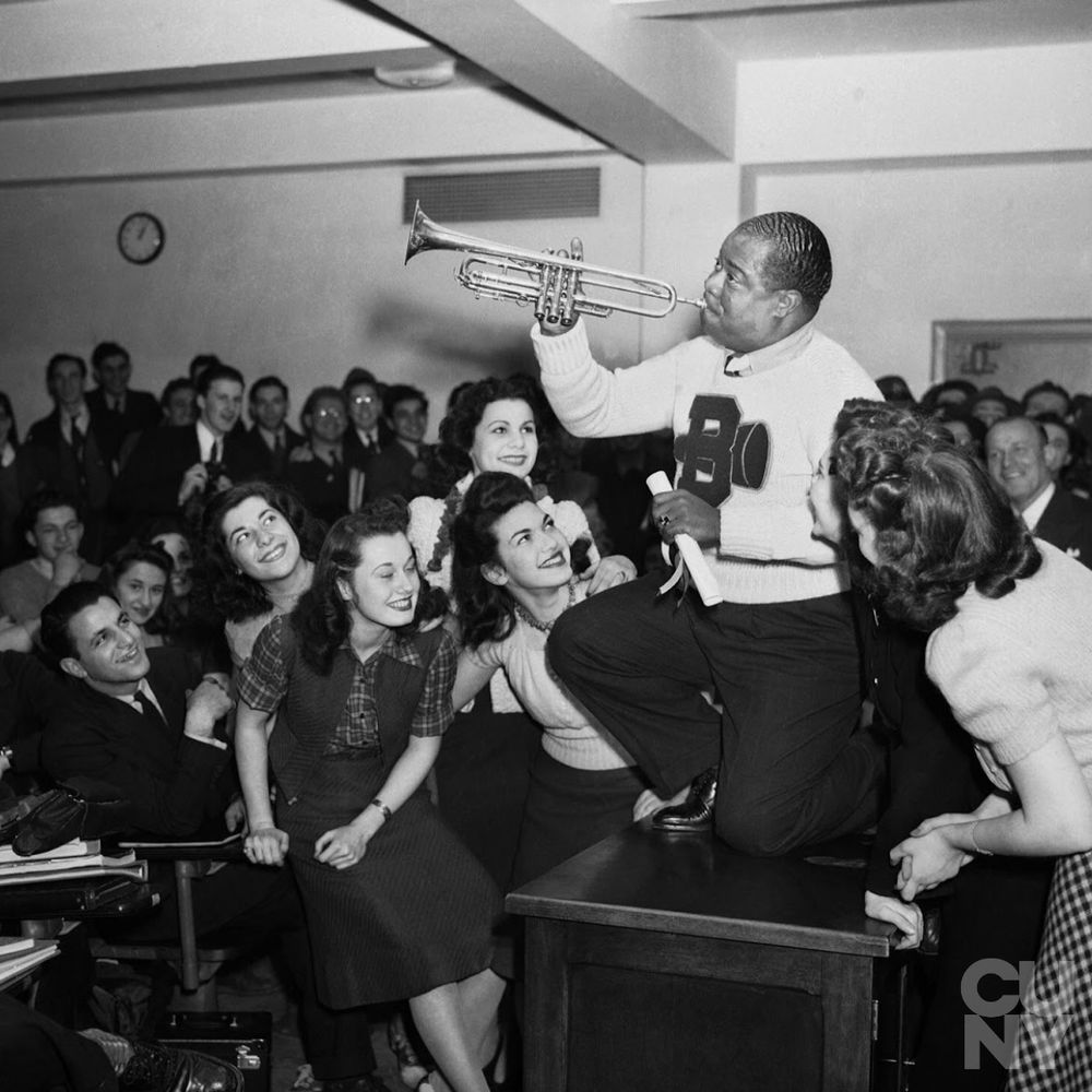 A black and white photograph shows Louis Armstrong standing on a desk in front of a crowded room at Brooklyn College. Armstrong is holding a trumpet and wearing a sweater. The audience, consisting of men and women, appears to be engaged.