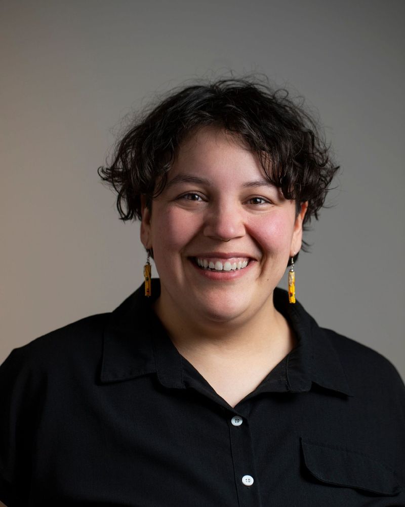 Photograph of Tyra Payer, a Native nonbinary person with short dark hair. In this photo they are wearing yellow earrings, a black button-down shirt, and a wide smile.