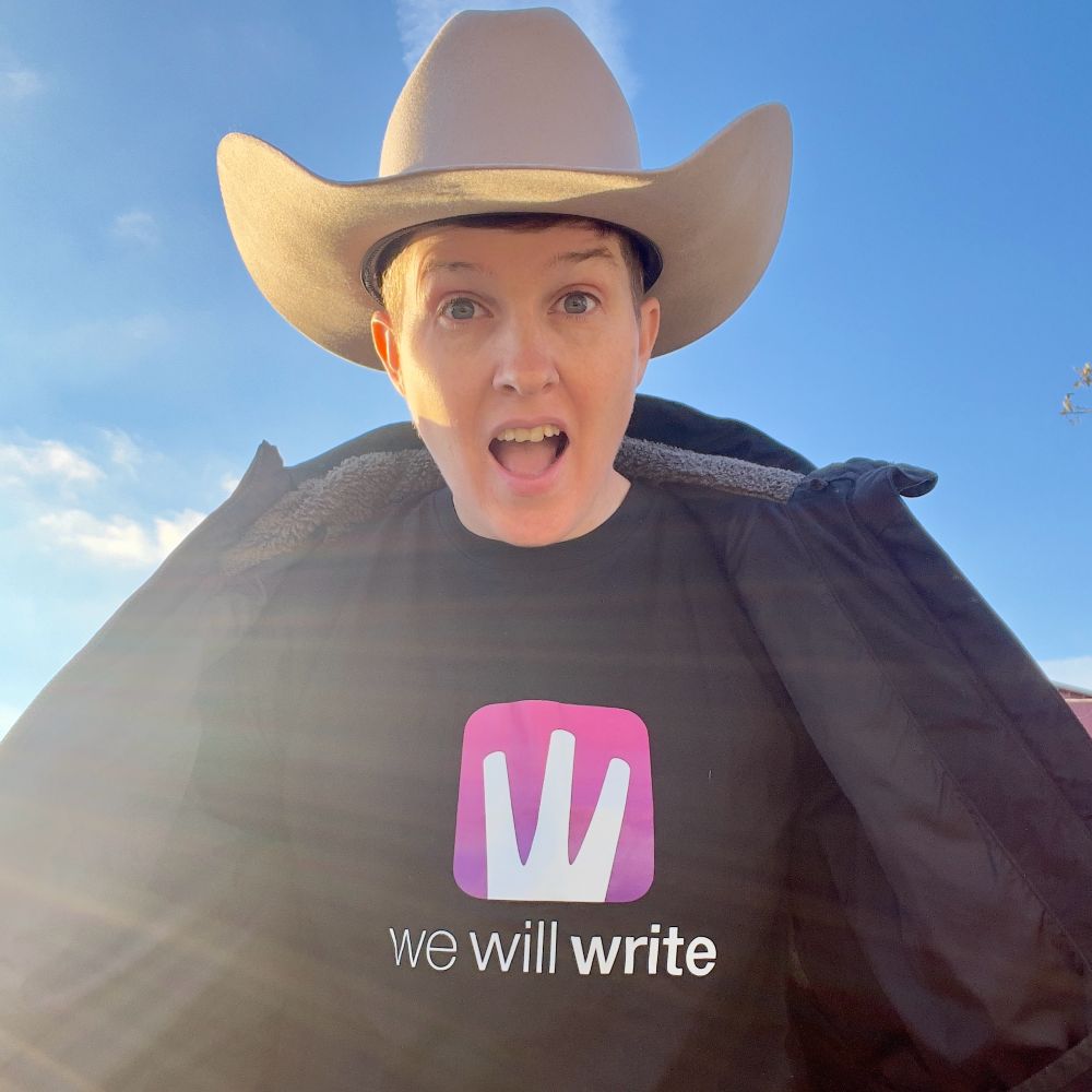 A person wearing a cowboy hat and a black T-shirt with the words “we will write” printed on it, along with a pink logo. They are outdoors under a clear blue sky, and sunlight is shining from the side, creating a glowing effect. Their expression is excited and surprised, with their mouth open and eyes wide. They are also wearing a jacket, which is open to reveal the T-shirt.