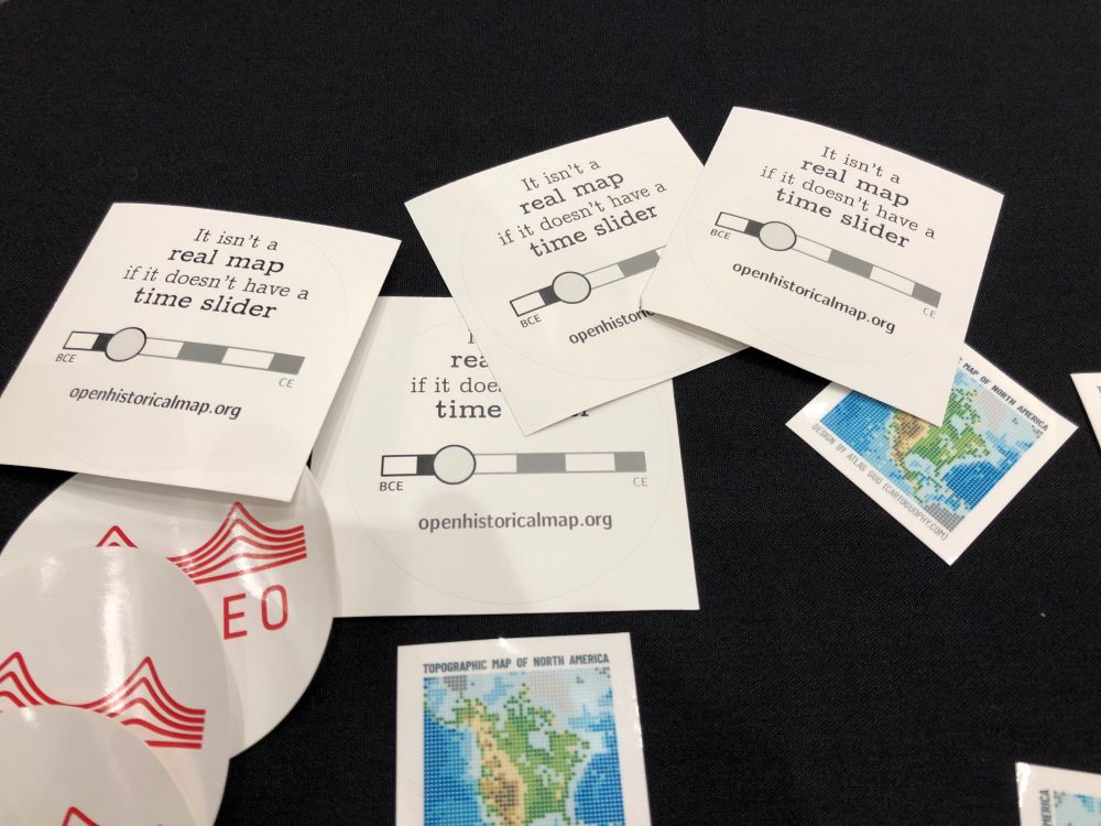 Four OpenHistoricalMap stickers say it isn’t a real map if it doesn’t have a time slider. The stickers are surrounded by other map-related stickers.
