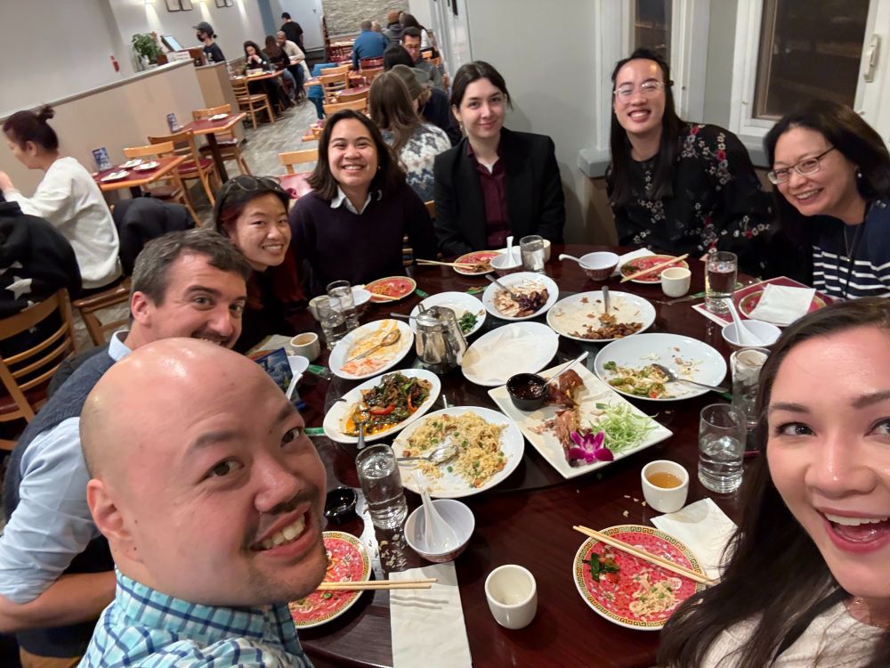 8 people smiling, sitting around a round table covered with plates of food