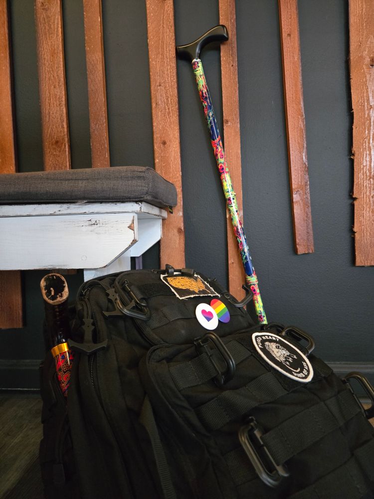 A 5.11 tactical bag and a cane leaning against a wall/bench. The cane features a colorful oil-slick appearance on the shft, and a black handle. The 5.11 tactical bag is black and features 2 patches on it: One with an opossum that reads "Pure of heart, dumb of ass", and another that has the US made out of bananas. Additionally, it has LGBTQ+ and bi pride pins, and a backup folding cane poking out of the side.