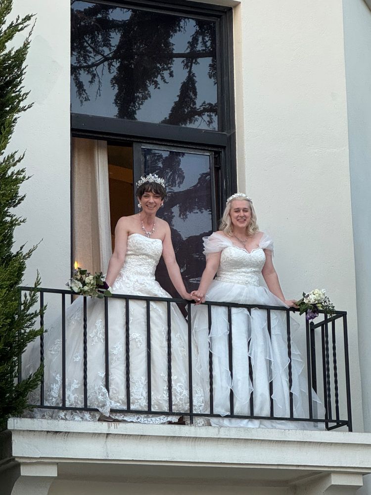 Kate and Rachael standing on a tiny balcony at Wrensmoor Castle about to throw bouquets to the crowd below. 