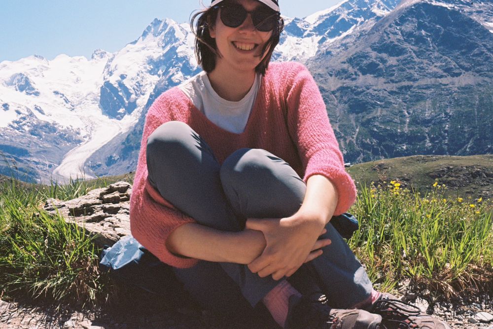 Amy, a tall, slender white woman, barely fitting in frame, wearing hiking clothes. She’s hunched over in the foreground, with the Bernina range in the background 