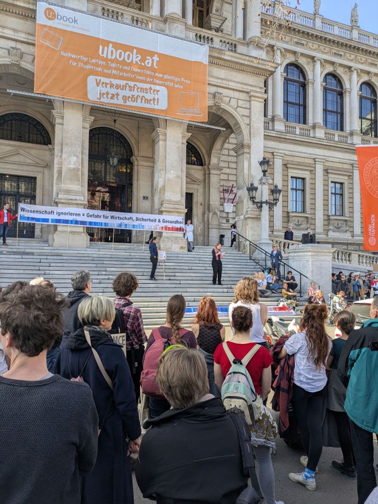 A group of people assembled in front of a regal building, which happens to be the main building of the University of Vienna, in Austria. 

They are listening to a woman speak as part of the 'March for Science' event. This event and the march that followed was in solidarity with the scientists in the USA. There is a climate change banner showing the increasing global temperature in the blue-red colorbar in the centre stage.