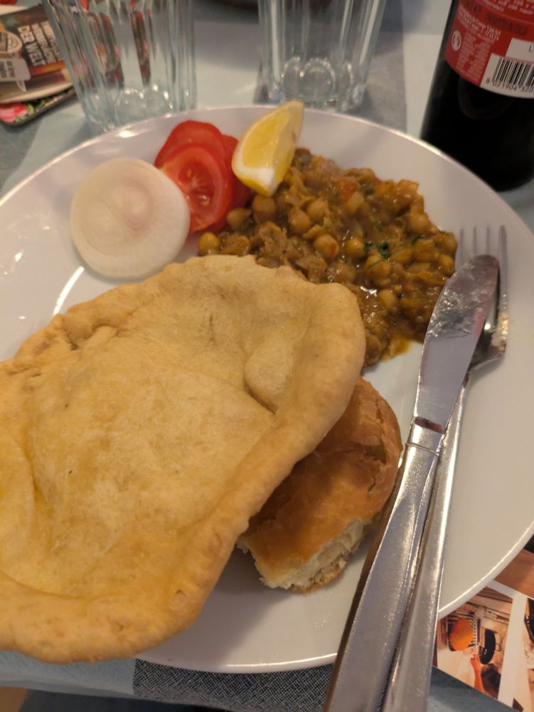 Shows a picture of food on a plate: with fried bread with some chickpeas curry, and a few slices of raw onion, tomato and lemon