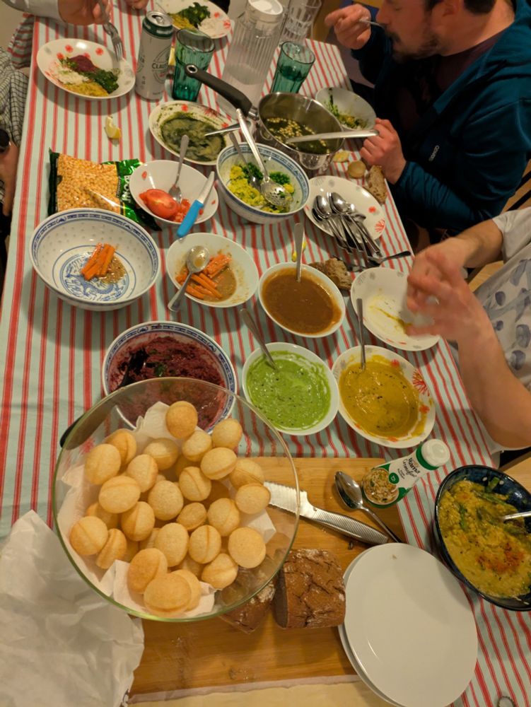 A table with various foods. Pani poori, chutneys, miso carrots, etc. On the side, a person is eating this