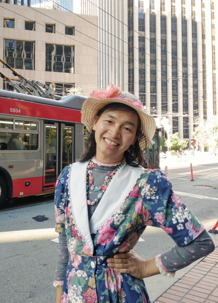 Alfred in downtown San Francisco, wearing a flowery hat and floral dress with a big collar. There is a trolleybus in the background 