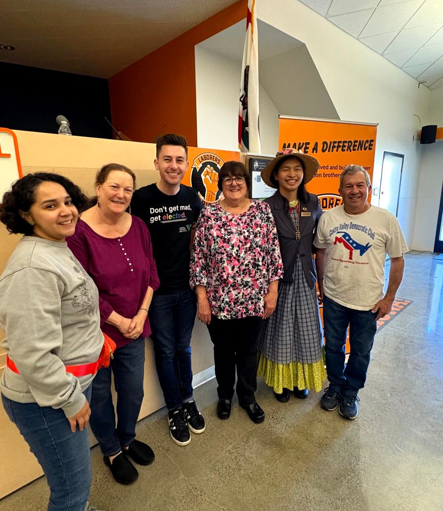 A group of six people standing in front of a California flag. One is wearing a shirt that says don't get mad, get elected, another one wearing a Castro valley Democrats shirt