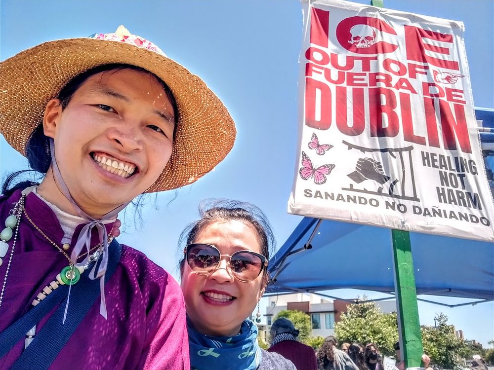 Alfred with another person in front of a Big cloth banner that says "ICE Out of Fuera de Dublin, Healing Not Harm, Sanando no Dañiando.  Banner has pictures of butterflies and two hands holding breaking out of a cage. 