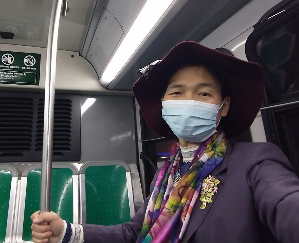 Alfred wearing a big hat, purple flowery scarf, gray jacket and long blue dress, on board an AC Transit bus with the famous bright green seats.
