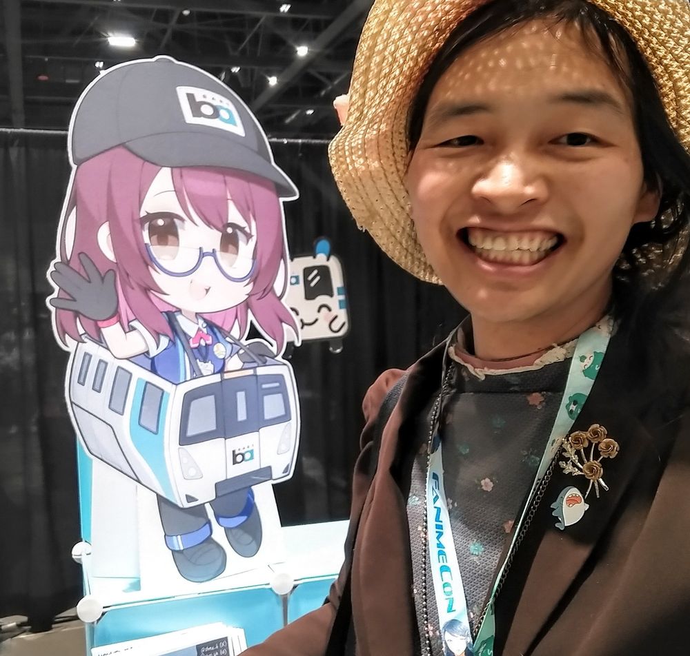 Alfred wearing a hat, a Fanime lanyard, and a brown blazer with pins next to a cutout of a BART anime character, Mira the train operator, wearing a bart train costume