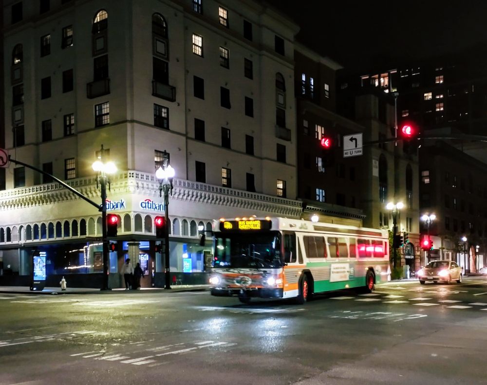 City street at night with an AC Transit bus in the classic orange and green and orange hubcaps