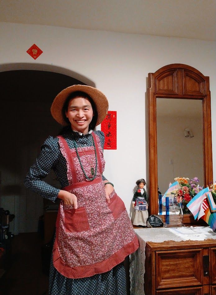 Alfred, wearing a straw hat, old-fashioned long sleeve green dress and red apron, and green beads and pendant, in front of a doorway. Above the door are a red diamond 福(luck) and to the right is a red vertical New Year banner 萬事如意 (may all things go well). At the right is a wooden cabinet with a large mirror above, there is a lace runner on the table, a doll wearing a Democratic Party apron, a couple of modern glass awards, and a basket of flags (parts of a trans flag, a US flag, a Progress Pride flag, and a nonbinary flag are visible) and fake flowers.