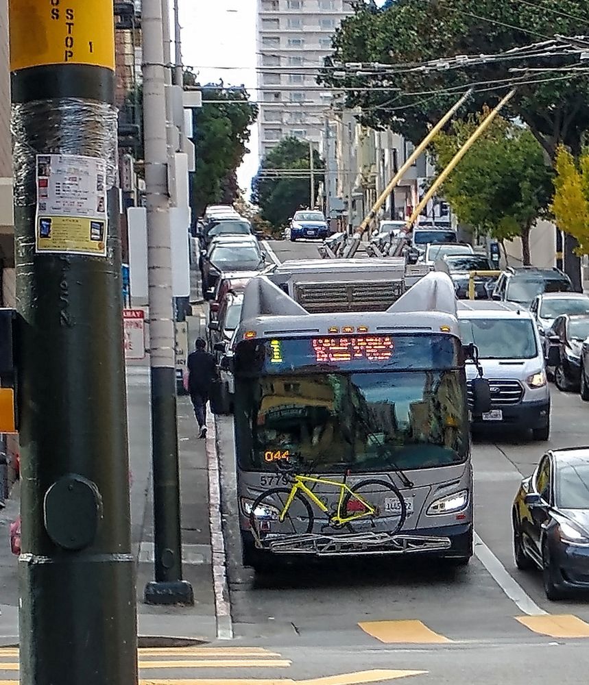 A 1-California trolley bus on a city street corner. The top of the bus has some streamlined design features that look like cat ears