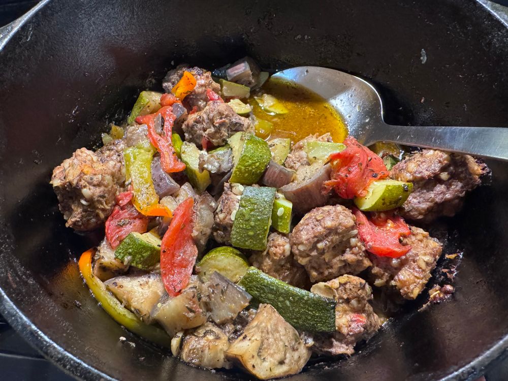Cast iron pot with metal spoon and mix of bright vegetables and dull meatballs. 