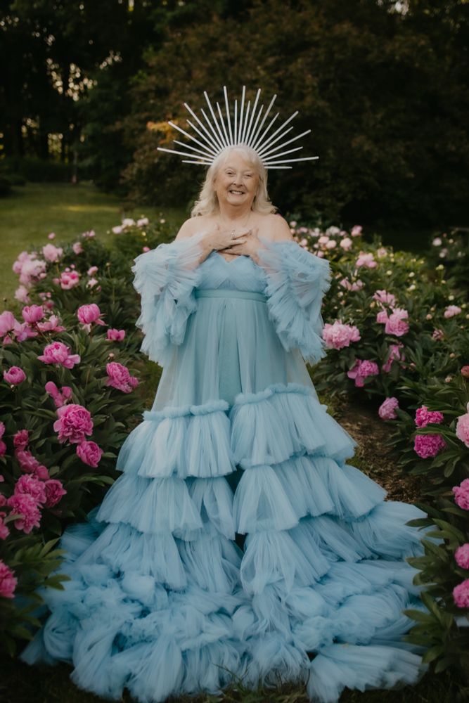 An older woman in a ruffled light blue ball gown, and spiked crown, surrounded by peonies