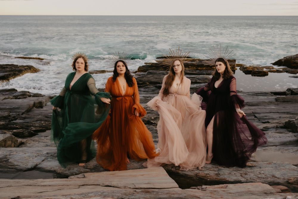 A group of four women, in colorful gowns, standing by the ocean 