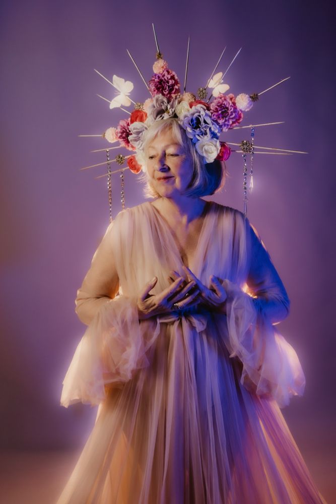 A photo of an older woman dressed in a flowing gown, with a spiked crown covered in pink and purple flowers and crystals