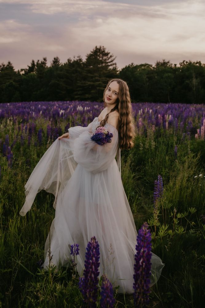 A woman with long hair in a silver gown, is standing in a field of purple and pink lupines 