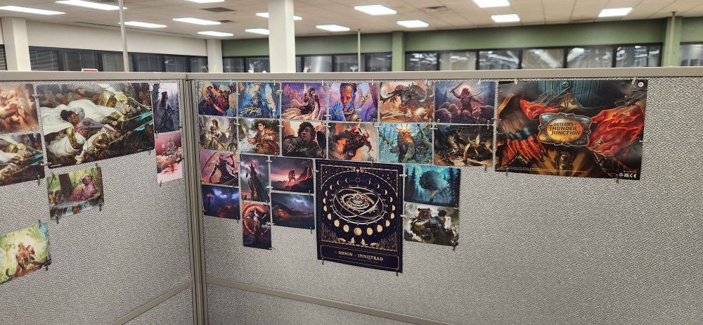 Magic the Gathering art cards span half the back wall of the office cubicle, showcasing some of the Lord of the Rings crossover cards, as well as beefy anthropomorphic characters that could trample OP, and he'd like it.