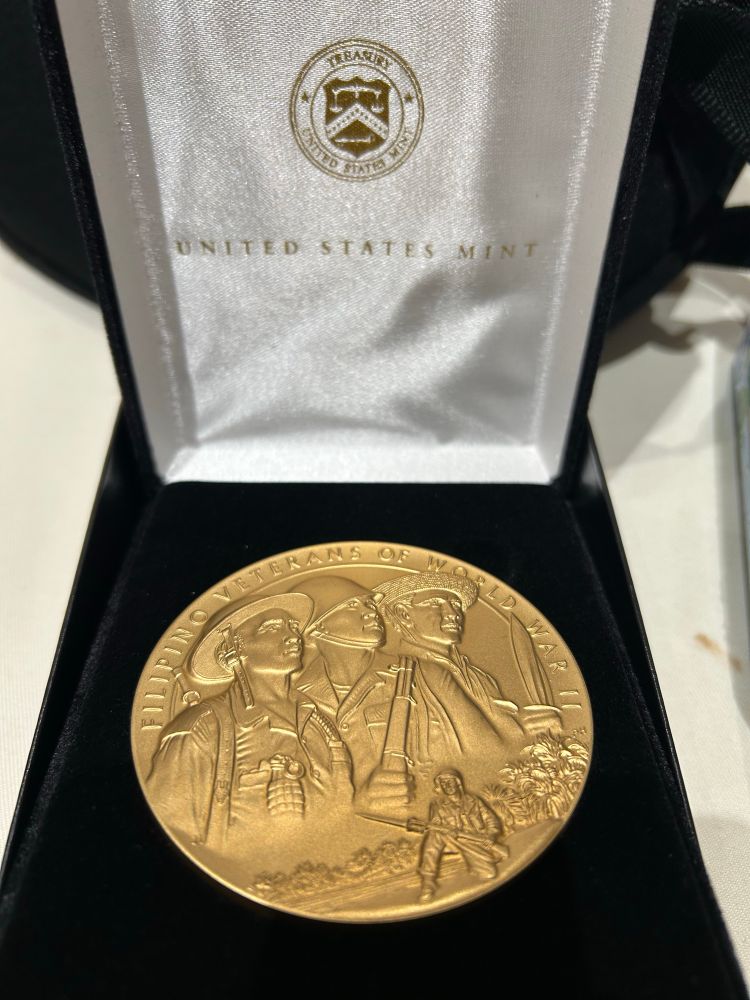A photo of a Congressional Gold Medal of Honor that specifically recognizes the Filipino veterans of WWII in a black velvet box with a white satin lid printed with “United States Mint.” The medal is embossed with the same design of Filipino fighters that appears on some US quarters. 
