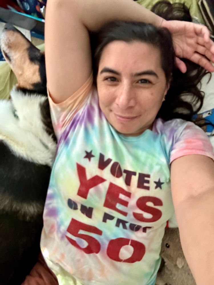 A brown person lying down, smiling up at the camera, next to a snoozing smooth-coat collie. They are wearing a tie-dye t-shirt with a stenciled message on it that says Vote yes on Prop. 50. 
