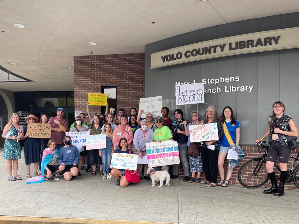 A group of people of different ages (teens to seniors) gathered beneath the Yolo County Library sign. They are holding signs decrying transphobia and bigotry. One kid is wearing a trans flag as a cape. 