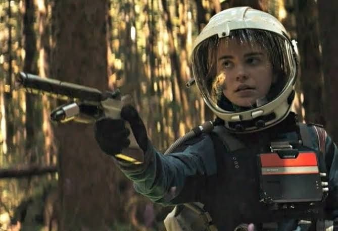 A girl in a retro-futuristic spacesuit wielding a pistol in a forest. Sophie Thatcher, from science-fiction film Prospect. 