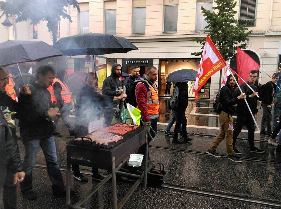 Dans cette image, nous voyons un groupe de manifestants défilant sous la pluie avec des parapluies et des drapeaux. Ce qui rend la scène particulièrement notable, c'est la présence d'un barbecue installé directement sur les rails du tramway. Les manifestants semblent profiter de cette surface stable pour griller de la viande en plein air, ajoutant une dimension conviviale et inattendue à leur protestation. L'arrière-plan montre des magasins, dont un "promod", et les manifestants portent des gilets de sécurité ainsi que des vêtements de pluie.