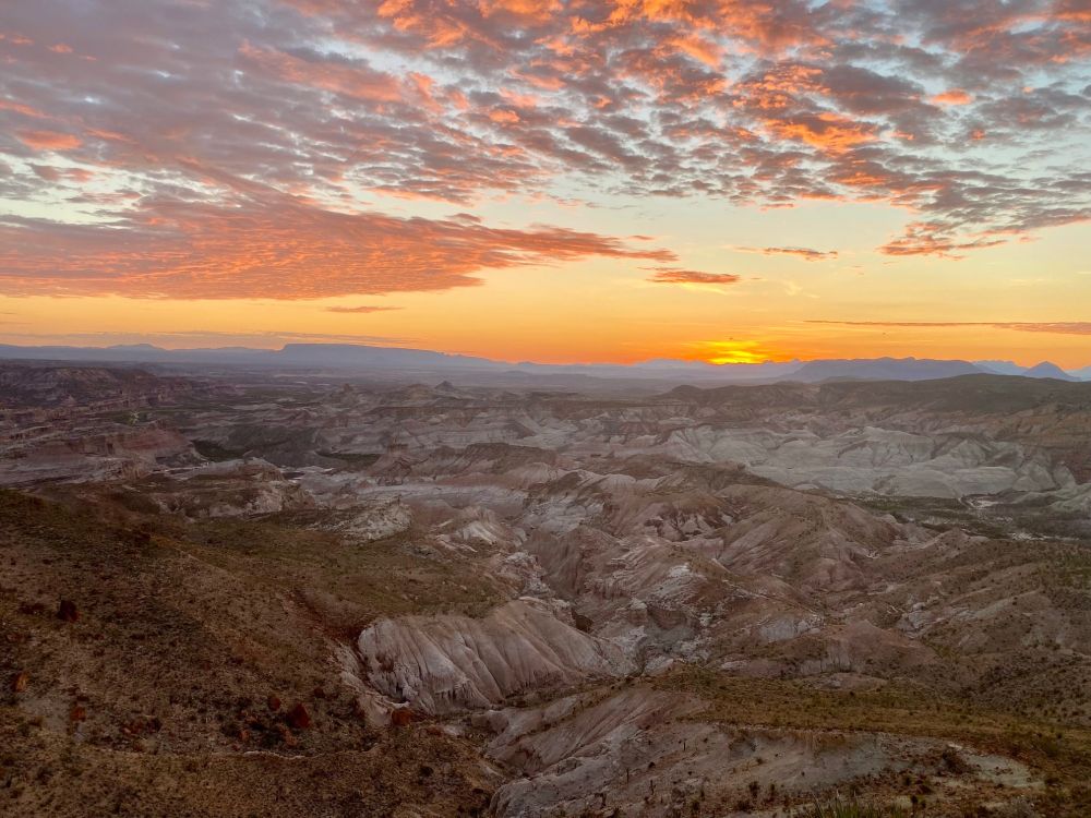 A morning view of the Devil's Graveyard. A bright, lurid sunrise peaks over the horizon; below, banded white and red cliffs roll in a badlands around a mostly dry creek canyon