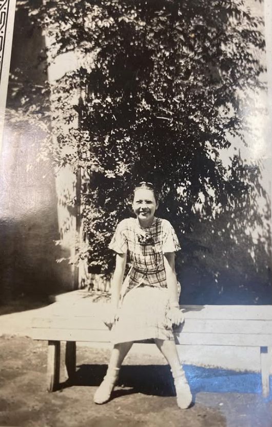A black and white photo of a young Maxine Langford in the 1930s at Texas Tech, smiling at the camera from a bench. She's wearing a knee-length dress, a plaid short sleeved shirt, and glasses. She's clearly very small. 