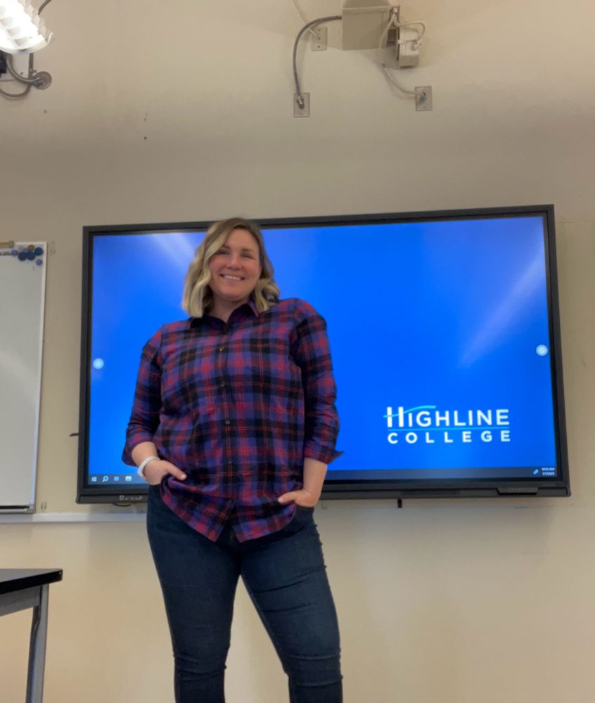 Tall blonde female teaching in front of classroom wearing a plaid flannel shirt because she thinks it strikes the right tone between scholarly and outdoorsy.