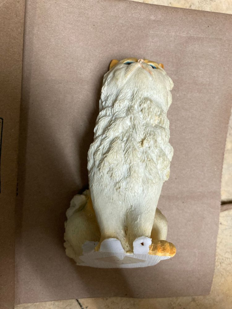 The front of a sitting Persian cat, off-white with pale flame point markings; three of her four paws are broken off. Fenris—a real black cat—is in the background 