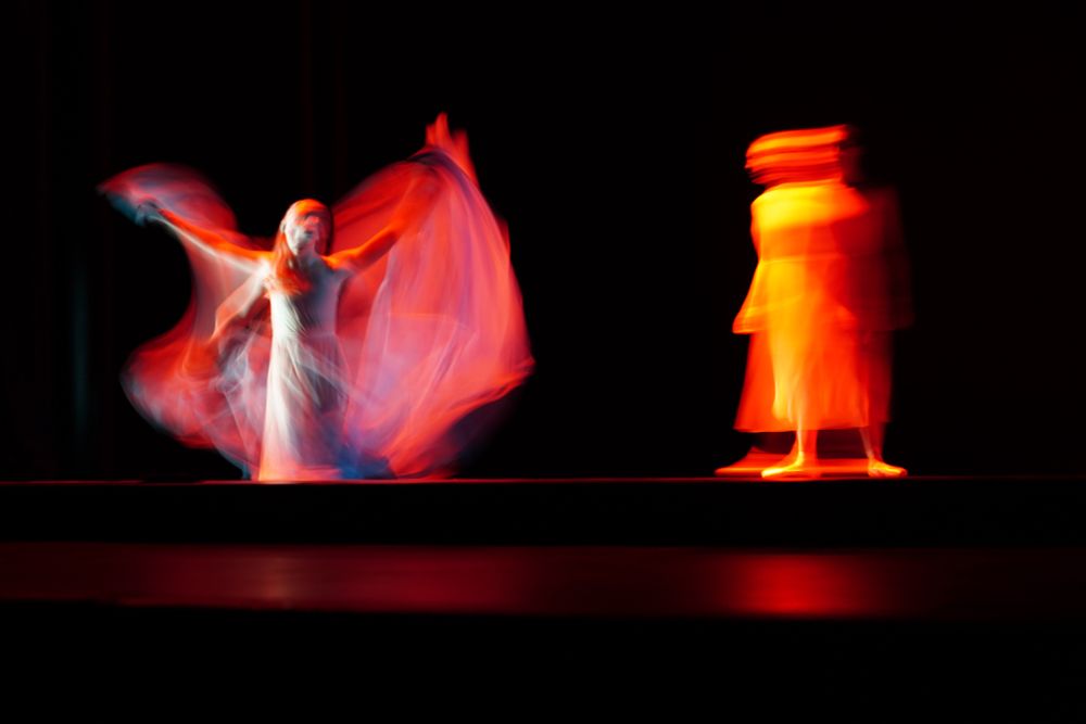 Two figures, very blurry. Look like ballerinas . One stands and looks on as another kneels and looks like she’s coming straight out of hell