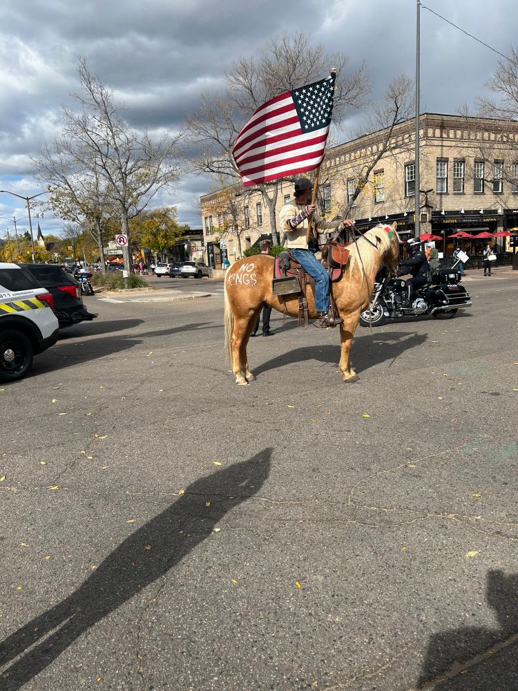 A man riding a palomino horse while holding an American flag. “No kings” is painted on the horses haunches.