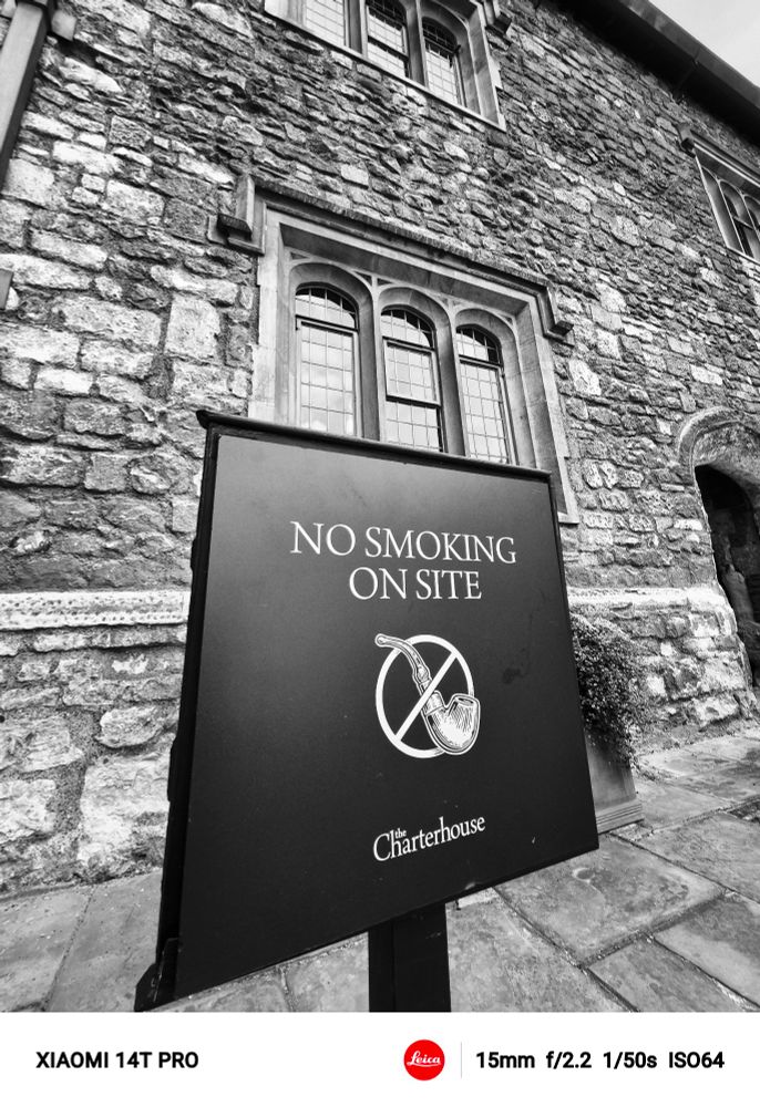 A sign with a pipe in a "no way" sign, with the title no smoking on site, and the name of the place, Charterhouse, with a background of a medieval monastery wall, in Central London 