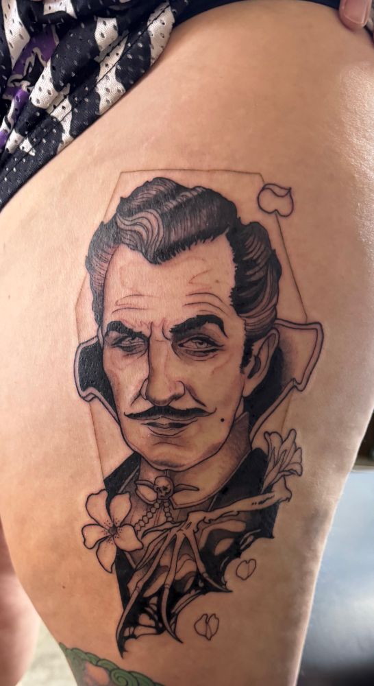A thigh tattoo of Vincent Price wearing Emmrich’s high collar and skull tie pin. There’s a bat wing, flower and coffin border. It’s grayscale.