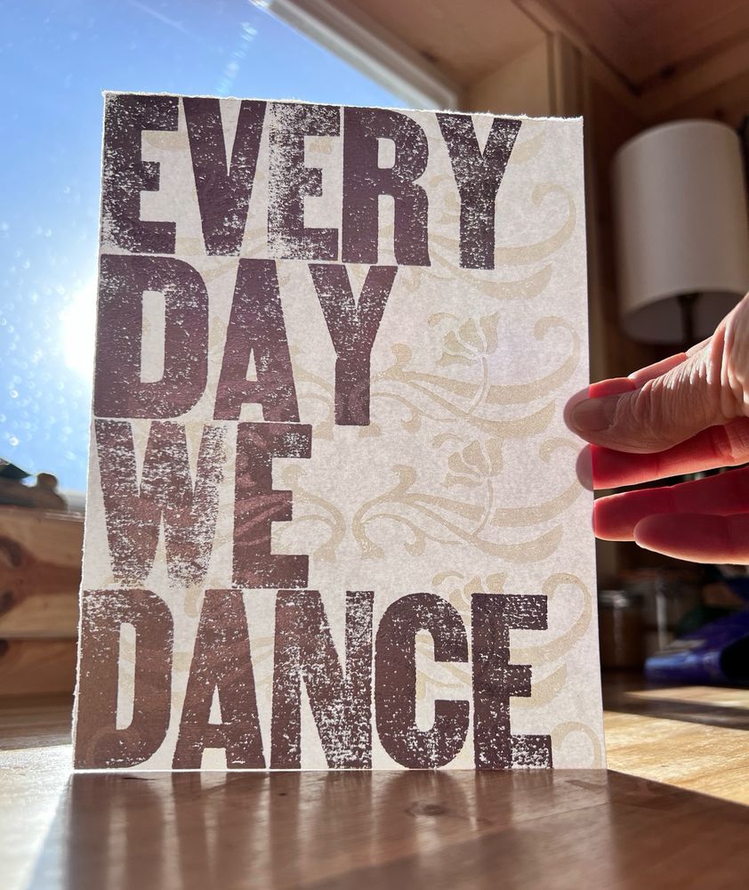 A letterpress print with the words ‘every day we dance’ printed in silver on a white background with white art nouveau-style flowers.