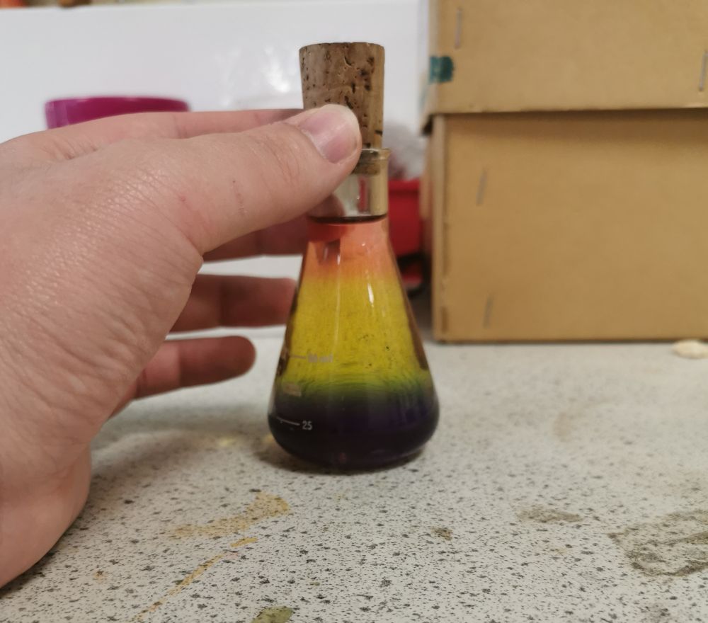 An image of a small conical glass bottle with a cork top. The liquid inside has a rainbow coloured gradient from top to bottom.